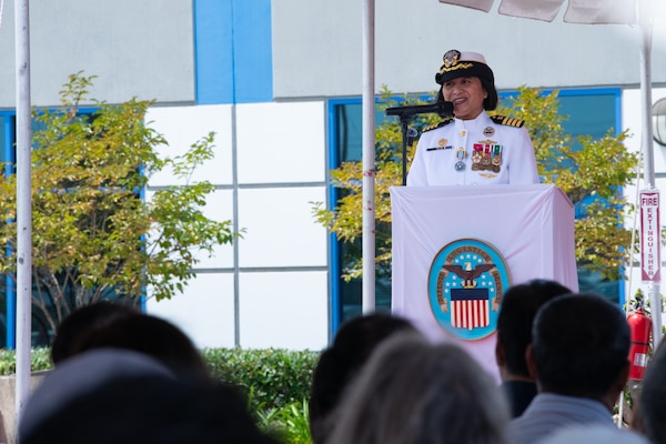 A woman wearing a military uniform stands behind a podium, outside on a stage under a tent, speaking to a crowd.