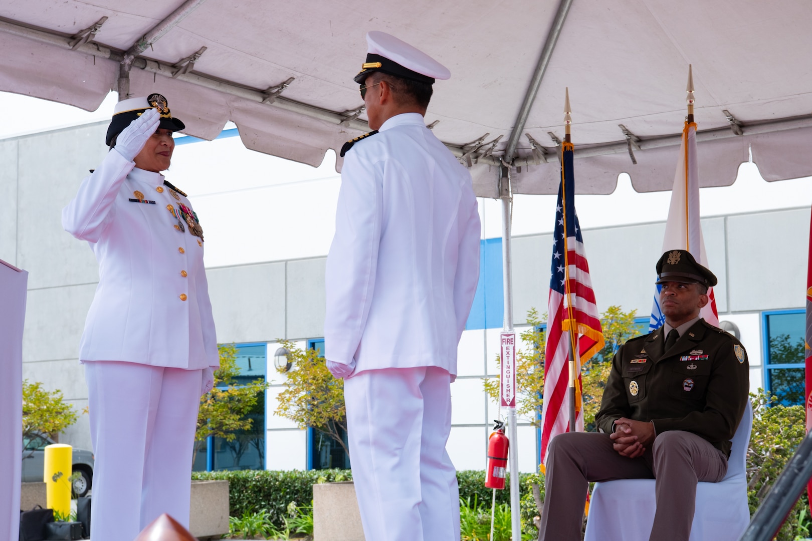A woman wearing a military uniform salutes a man wearing a military uniform while standing outside on a stage covered by a tent.