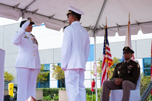 A woman wearing a military uniform salutes a man wearing a military uniform while standing outside on a stage covered by a tent.