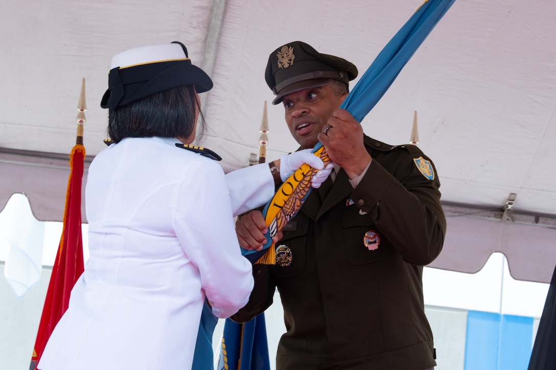 A woman wearing a military uniform passes a flag to a man wearing a military uniform on a stage under a tent outside.