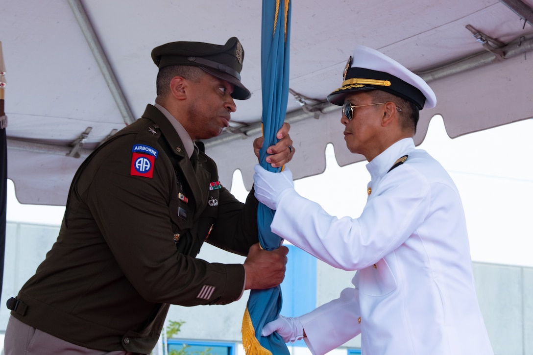 A man wearing a military uniform passes a flag to another man wearing a military uniform on a stage outside under a tent.