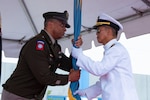 A man wearing a military uniform passes a flag to another man wearing a military uniform on a stage outside under a tent.