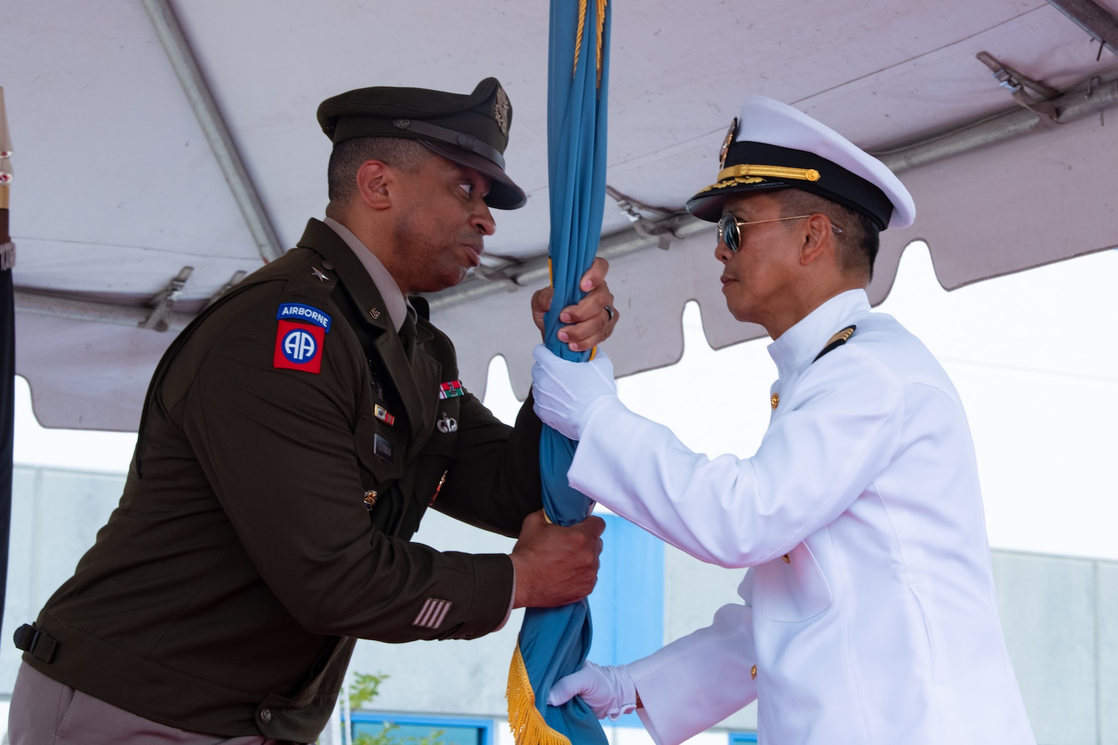 A man wearing a military uniform passes a flag to another man wearing a military uniform on a stage outside under a tent.