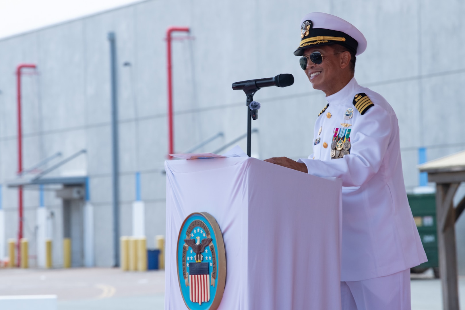 A man wearing a military uniform stands behind a podium, outside on a stage under a tent, speaking to a crowd.