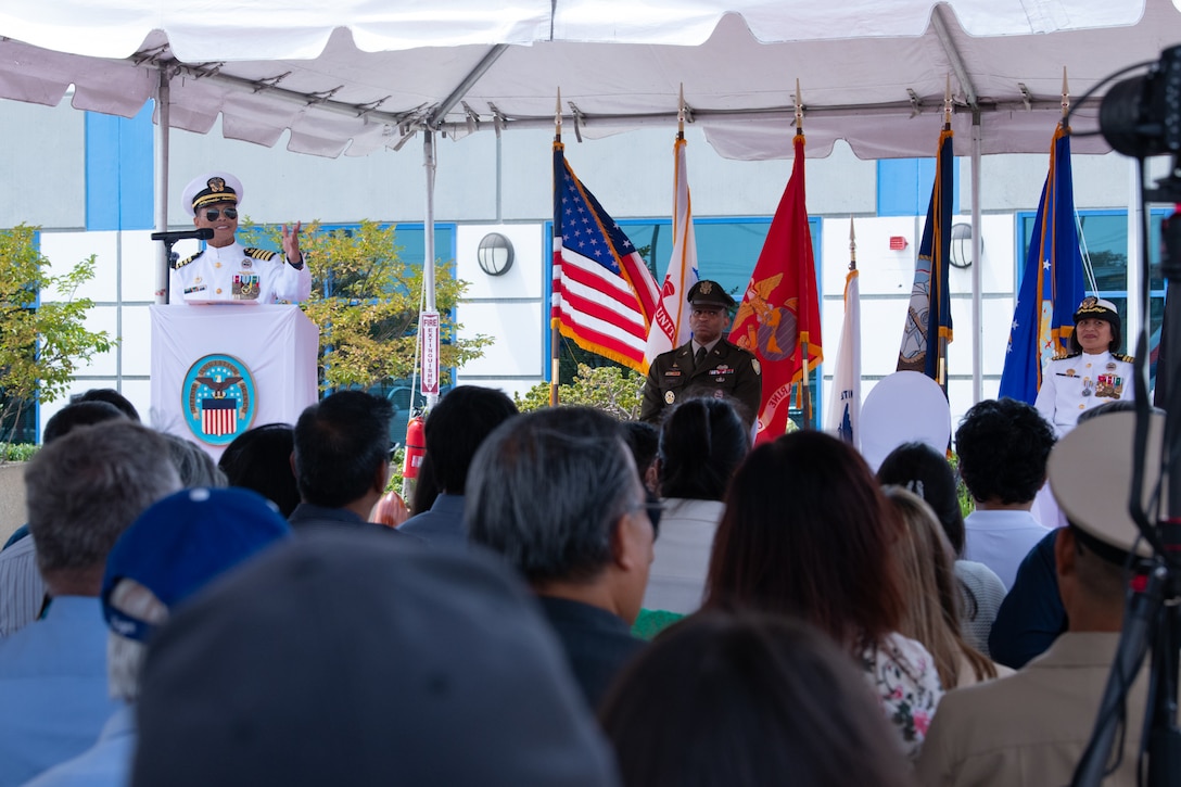 A man wearing a military uniform stands behind a podium, outside on a stage under a tent, speaking to a crowd.
