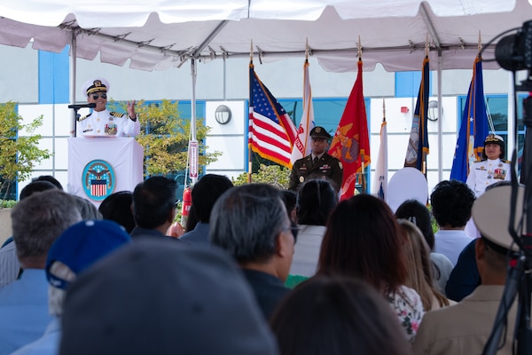 A man wearing a military uniform stands behind a podium, outside on a stage under a tent, speaking to a crowd.