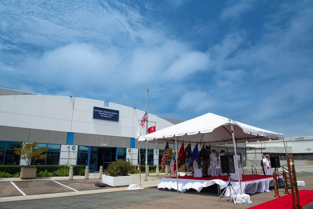 People stand on a stage under a tent in front of a building outside on a sunny day.