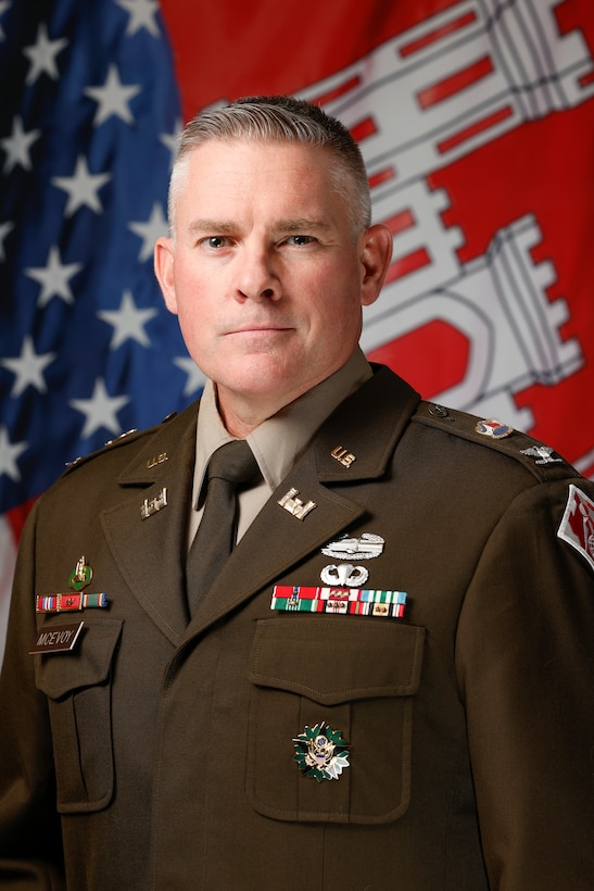 A formal head-and-shoulders portrait of a middle-aged white man in a U.S. Army Service Uniform. He has short, graying hair and a serious expression, looking directly at the camera. His olive-brown jacket is decorated with various medals, ribbons, and insignia, including a name tag that reads "McEVOY". Behind him, an American flag is visible on the left, and a red flag with the white U.S. Army Corps of Engineers castle emblem is on the right.