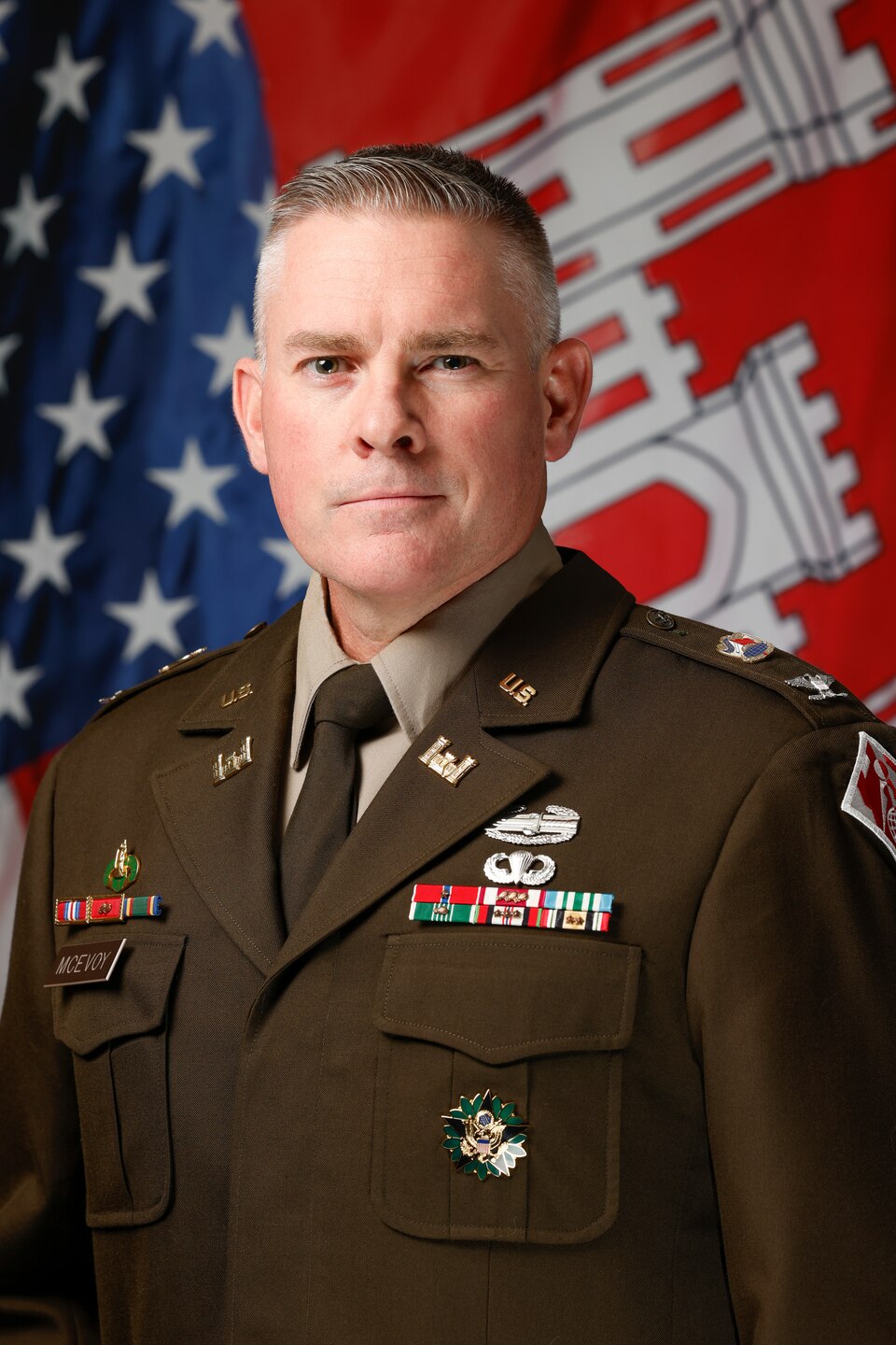 A formal head-and-shoulders portrait of a middle-aged white man in a U.S. Army Service Uniform. He has short, graying hair and a serious expression, looking directly at the camera. His olive-brown jacket is decorated with various medals, ribbons, and insignia, including a name tag that reads "McEVOY". Behind him, an American flag is visible on the left, and a red flag with the white U.S. Army Corps of Engineers castle emblem is on the right.