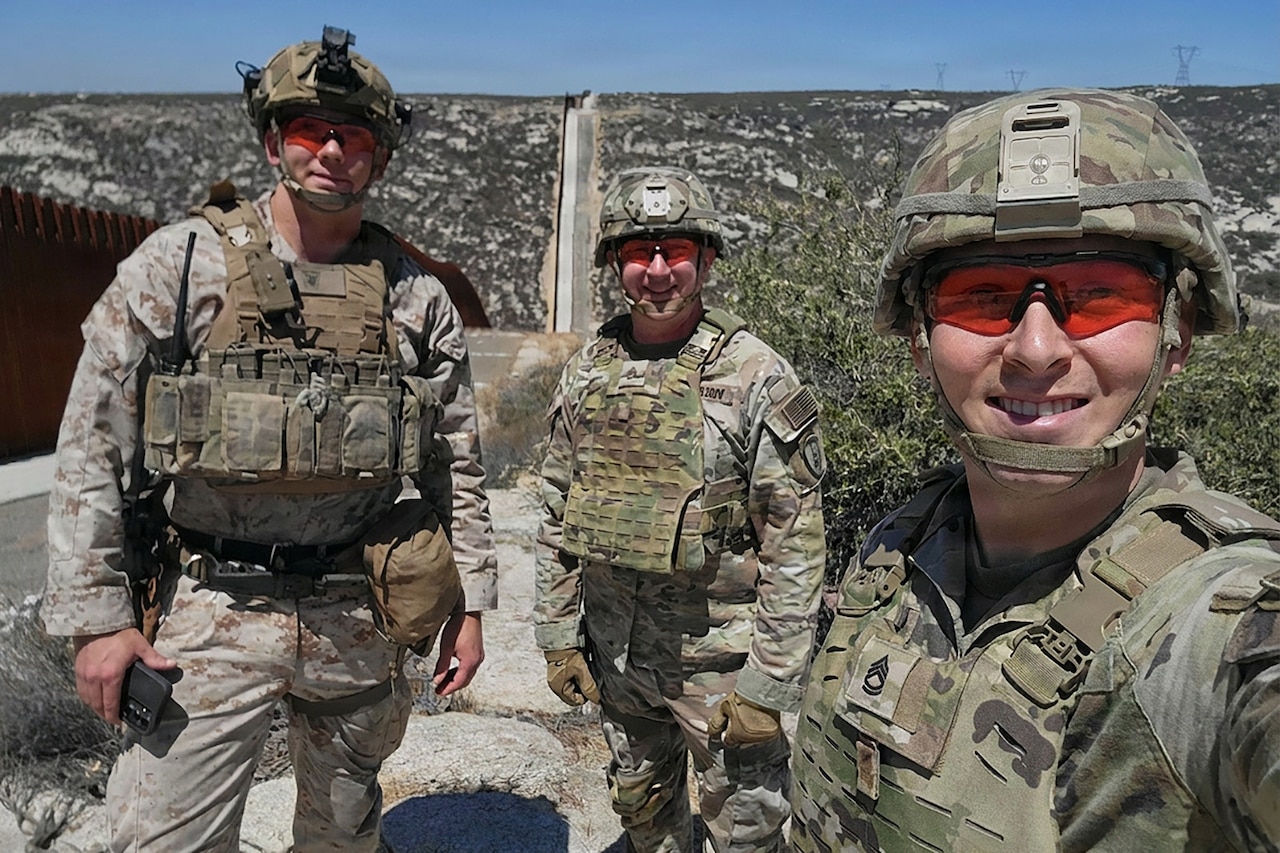 Three men wearing camouflage military uniforms, helmets and sunglasses smile as they pose for a photo in the desert.