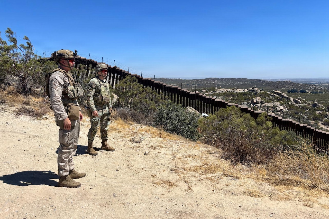 Two men wearing camouflage military uniforms, helmets and sunglasses survey a border fence in the desert.