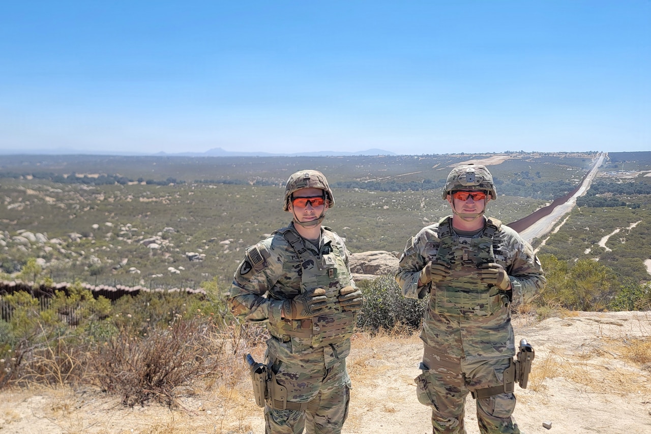 Two men wearing camouflage military uniforms, helmets and sunglasses smile as they pose for a photo in the desert.