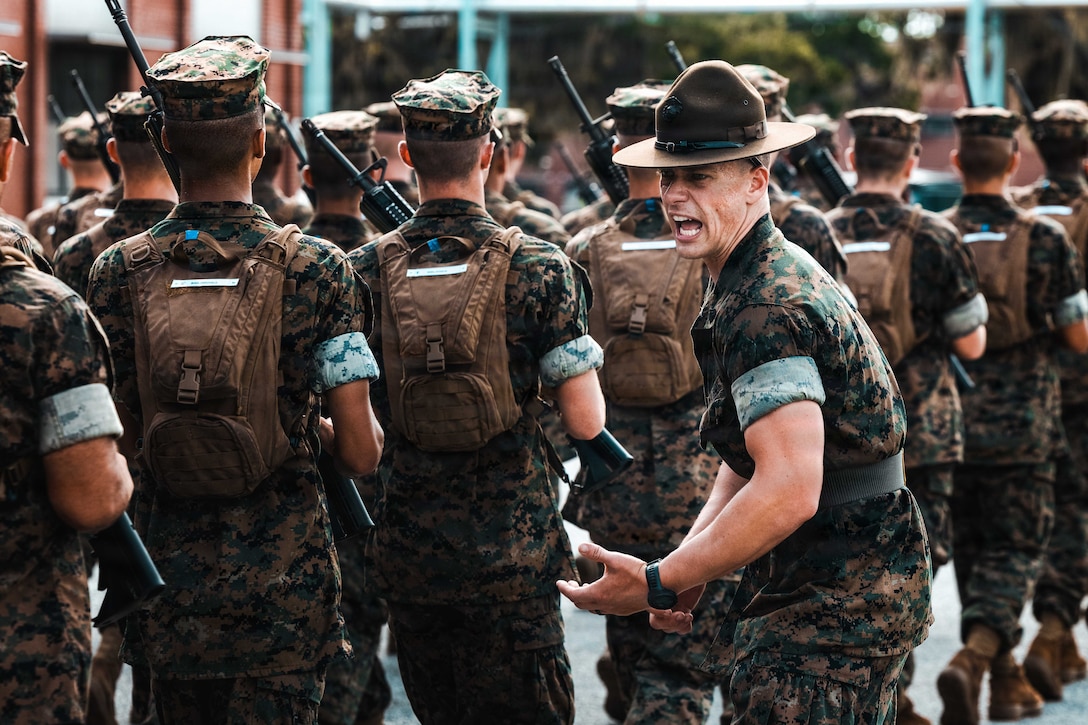 A Marine yells at a group of Marine recruits walking in formation with rifles and backpacks.