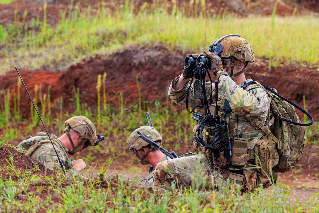 Two soldiers sit in a trench and another kneels on the ground slightly above them and uses binoculars.