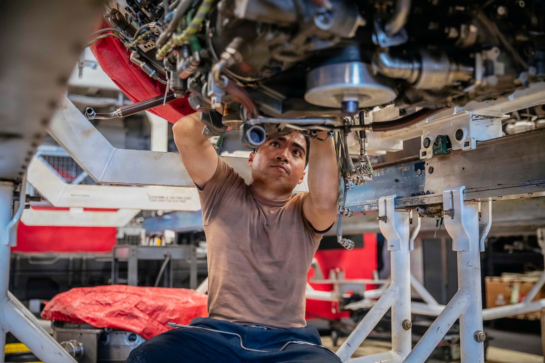 A sailor in a brown shirt works on an engine in a maintenance room aboard a ship.
