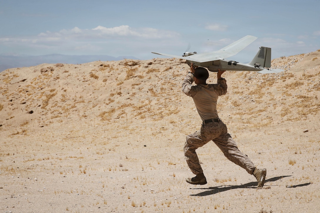 A Marine runs in a desert-like area while preparing to launch an RQ-20 Puma into a blue sky.