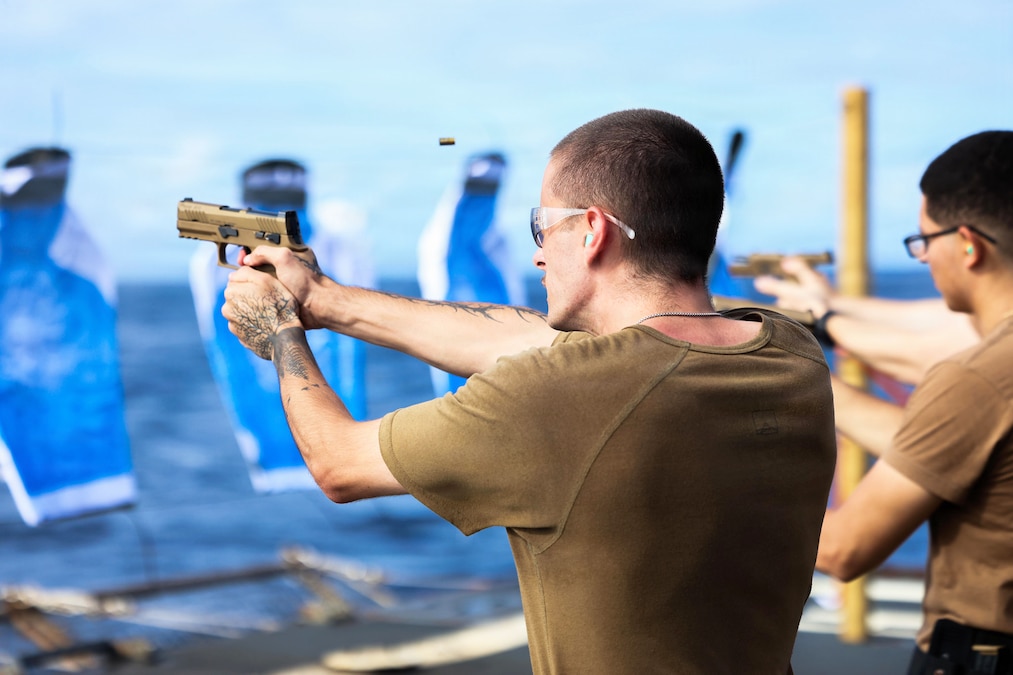 Ammo flies in the air as sailors wearing protective eyewear fire weapons toward blue targets aboard a ship at sea.