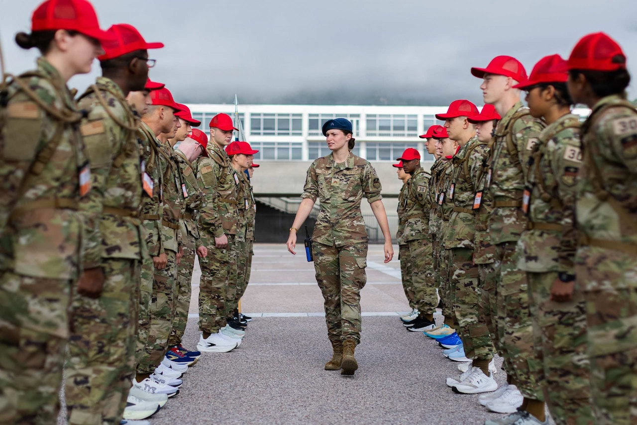 A woman wearing a camouflage military uniform and a blue beret walks between a formation of people in camouflage military uniforms and red hats.
