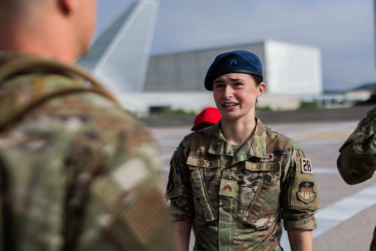 A woman wearing a camouflage military uniform and a blue beret looks at another person in similar attire.
