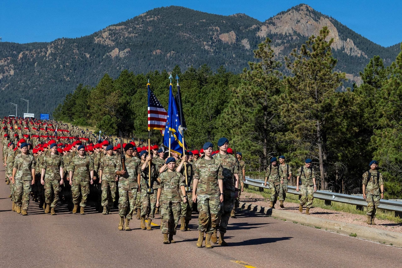 A group of people wearing camouflage military uniforms and red and blue berets march down a road in formation. A forest and mountain range is behind them.