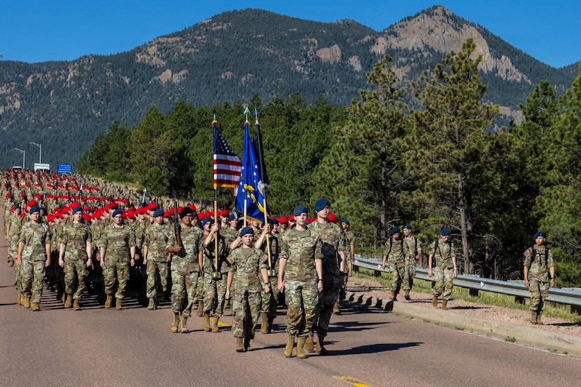 A group of people wearing camouflage military uniforms and red and blue berets march down a road in formation. A forest and mountain range is behind them.