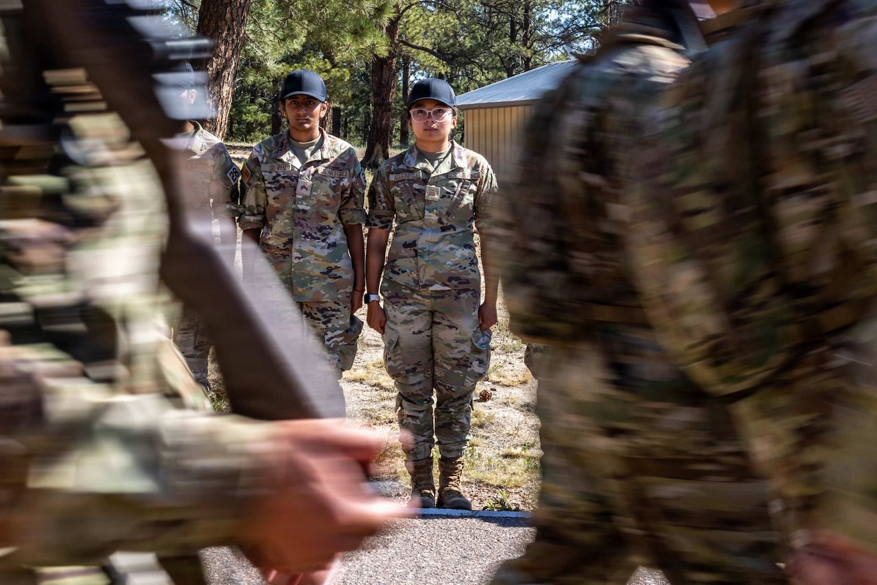 Two people wearing camouflage military uniforms and black hats stand at attention as a formation marches by.