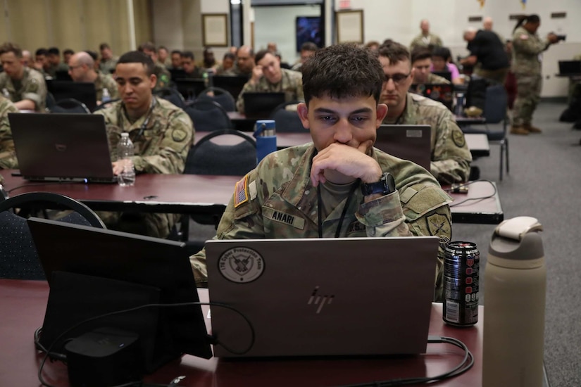 man wearing a camouflage military uniform sits at a table and looks at a computer. Numerous other men and women in similar attire do the same in the background.
