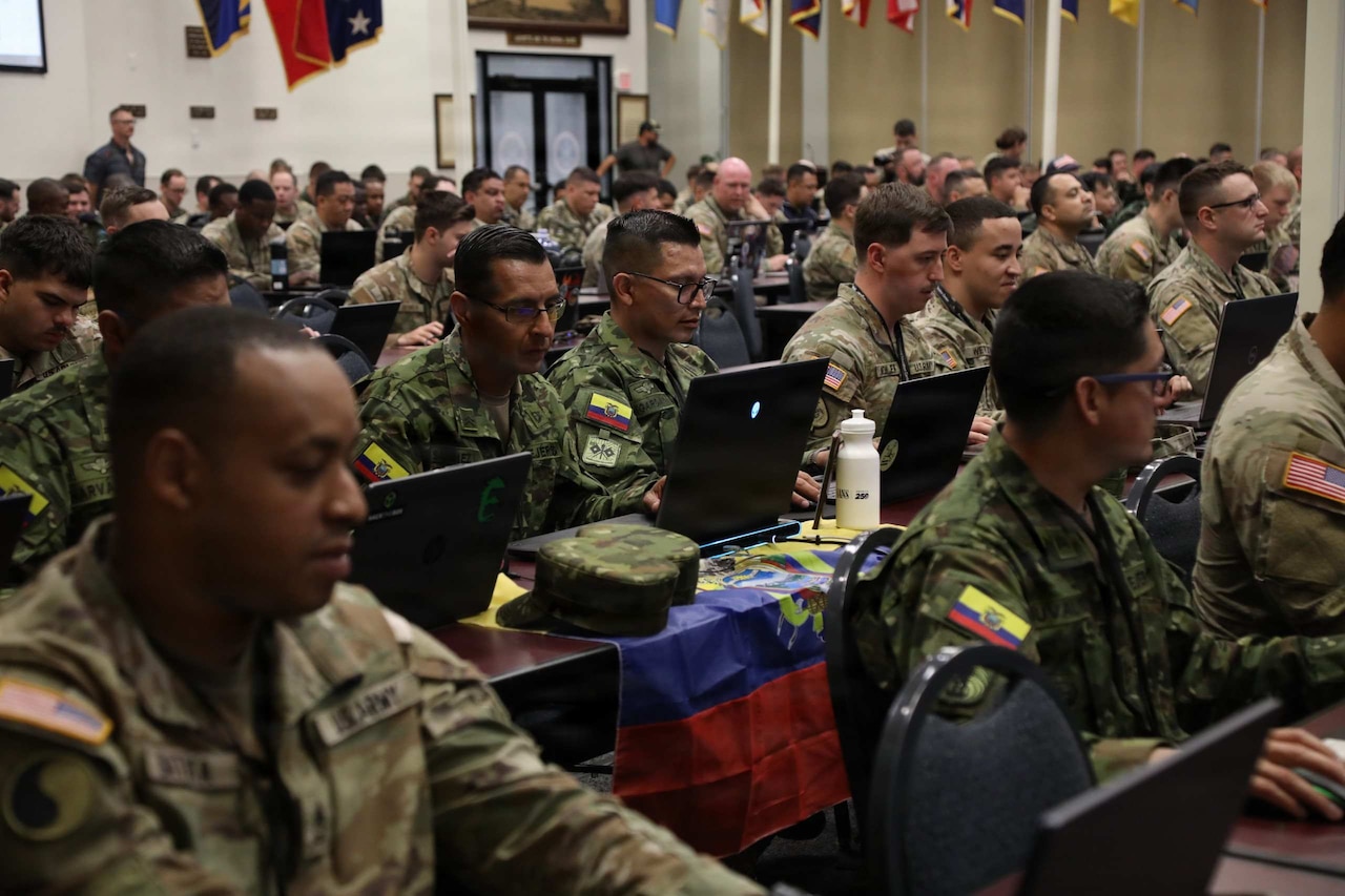 Dozens of people in various styles of camouflage military uniforms sit at tables in a large room working on computers.