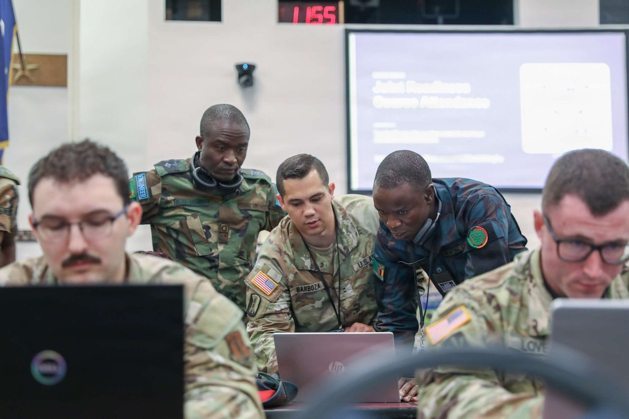 Three men wearing camouflage military uniforms stand and look at a computer in a large room.