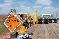 An explosives warning placard sits in focus on a trailer as U.S. Air Force Airmen assigned to the 7th Munitions Squadron load munitions onto the trailer bed using a forklift during the Death Strike: Trident Fury at Dyess Air Force Base, Texas, June 22, 2026. Death Strike: Trident Fury is a multi-day munitions production exercise focused on strengthening interoperability and proving the joint force's ability to rapidly scale operations in a simulated combat environment. (U.S. Air Force photo by Airman 1st Class William Neal)