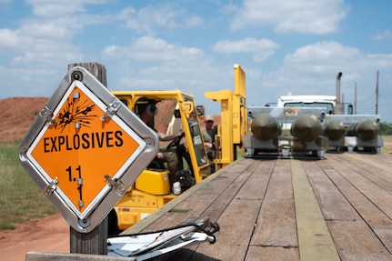 An explosives warning placard sits in focus on a trailer as U.S. Air Force Airmen assigned to the 7th Munitions Squadron load munitions onto the trailer bed using a forklift during the Death Strike: Trident Fury at Dyess Air Force Base, Texas, June 22, 2026. Death Strike: Trident Fury is a multi-day munitions production exercise focused on strengthening interoperability and proving the joint force's ability to rapidly scale operations in a simulated combat environment. (U.S. Air Force photo by Airman 1st Class William Neal)