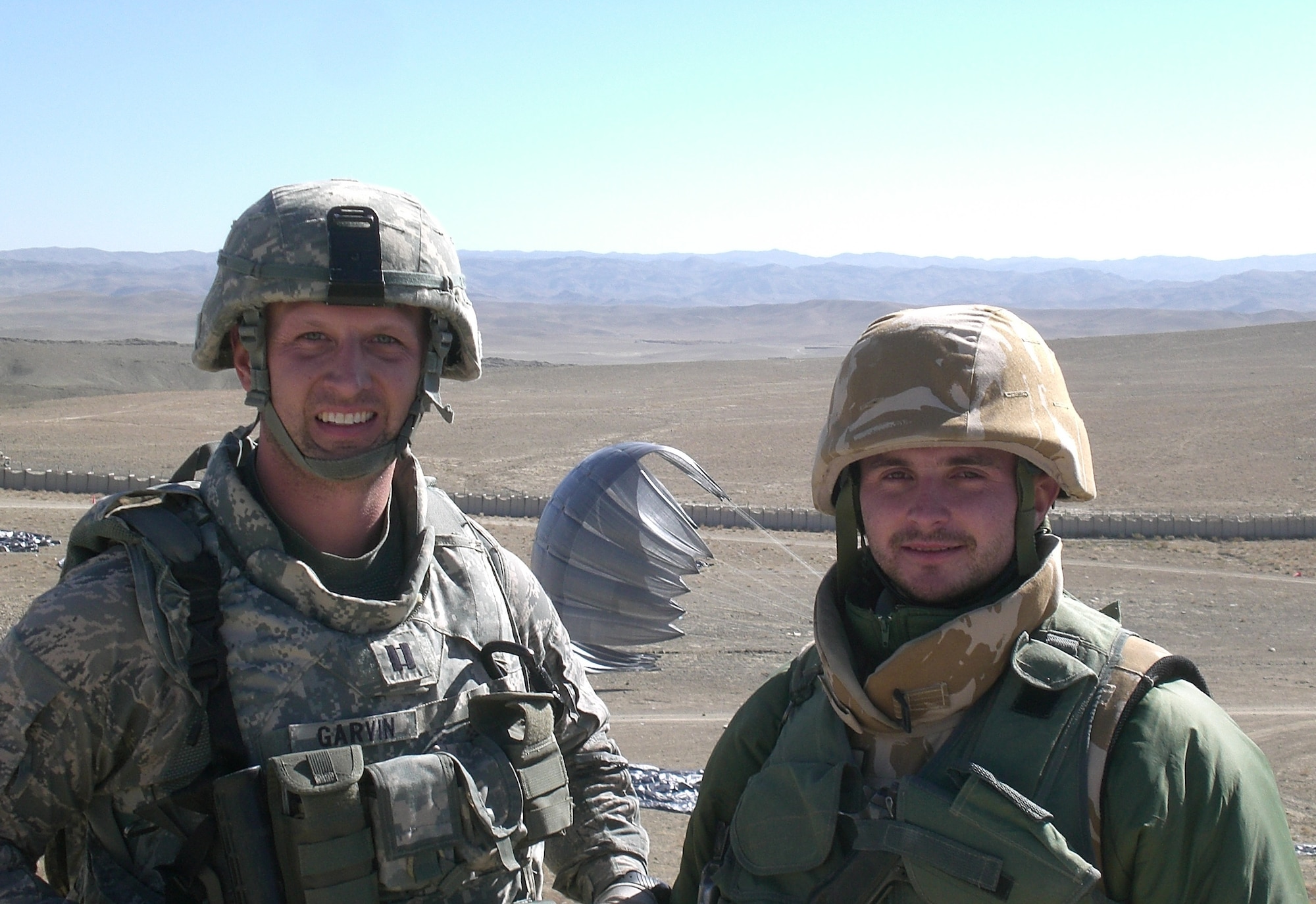 Two military members stand in a desert with a parachute in the background.