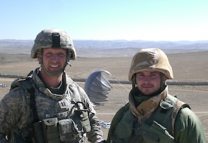 Two military members stand in a desert with a parachute in the background.