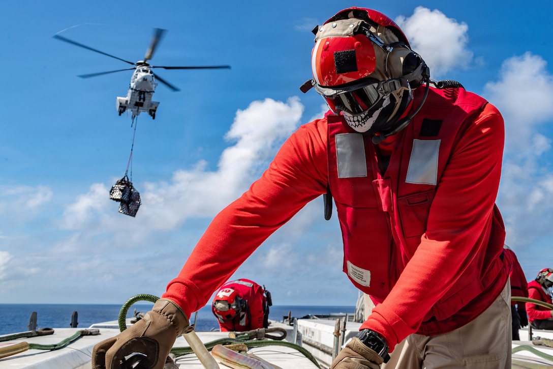 A person in a red shirt and helmet wears dark goggles over a face mask with teeth painted on it while on a ship's deck. Cargo hanging from a helicopter is in the background.