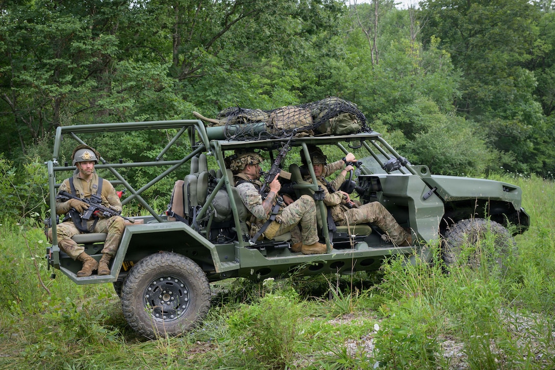 Soldiers ride in a vehicle without doors, with some holding rifles, over rocky and weed-covered terrain. Bags in a net sit on the roof.