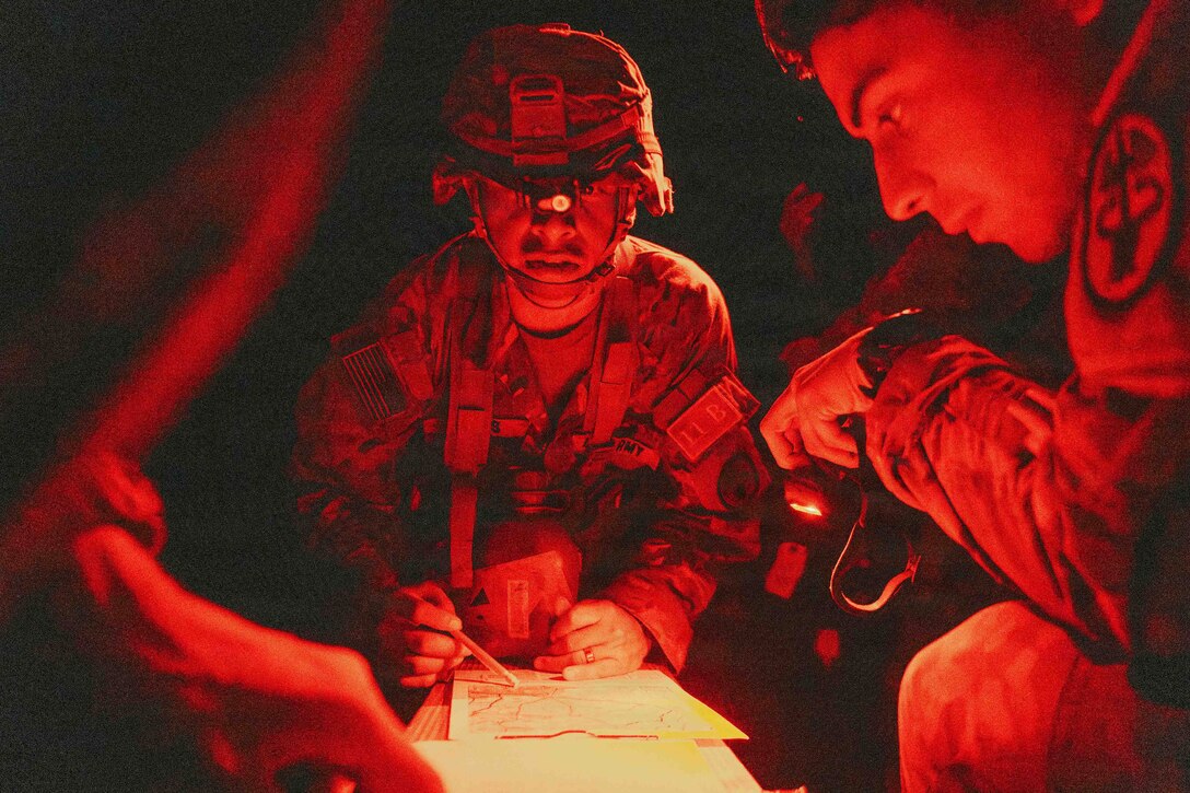 Red light is reflected on a soldier holding a pencil over a map at night. Other soldiers sit nearby.