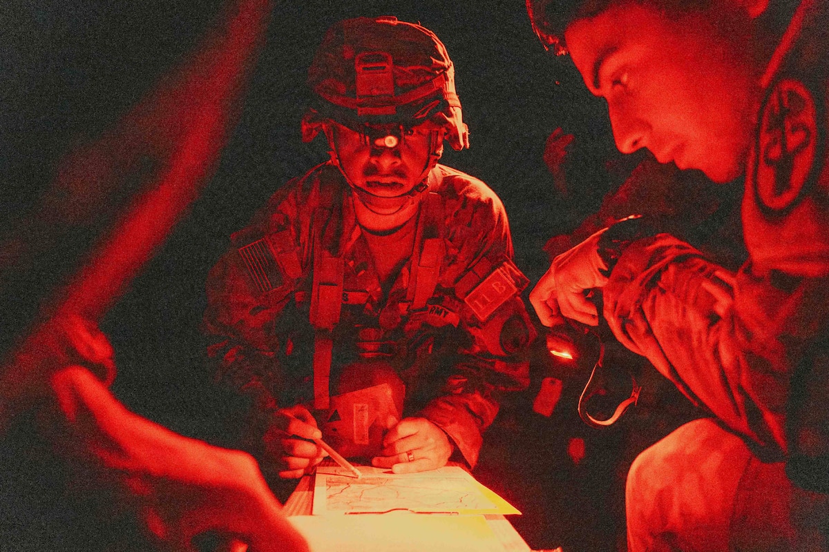 Red light is reflected on a soldier holding a pencil over a map at night. Other soldiers sit nearby.