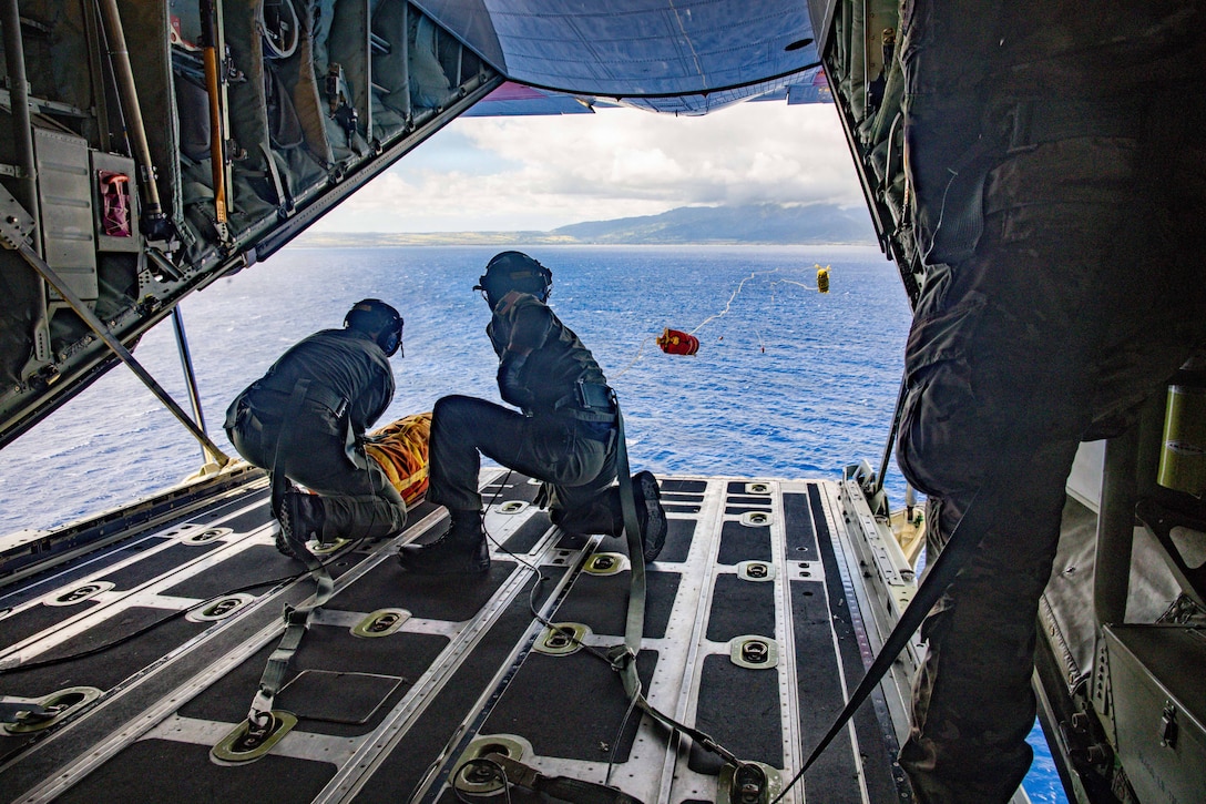 Two service members face the ocean from the open cargo port of an aircraft in flight and throw floatation devices connected to rope toward the water.