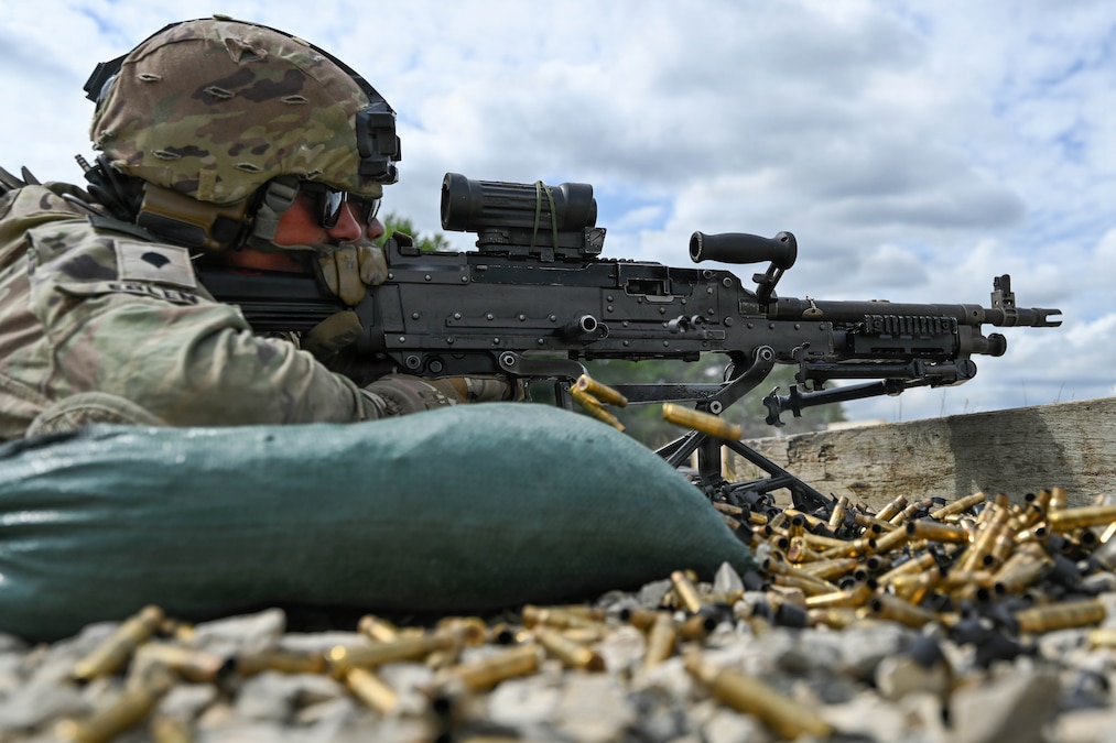 A soldier lies on the ground and fires a machine gun. Shell casings are in the air and in piles on the ground. Another soldier is in the background.