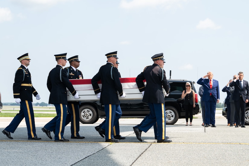 Six men in formal military uniforms carry a flag-draped case as people in similar attire salute in the background.