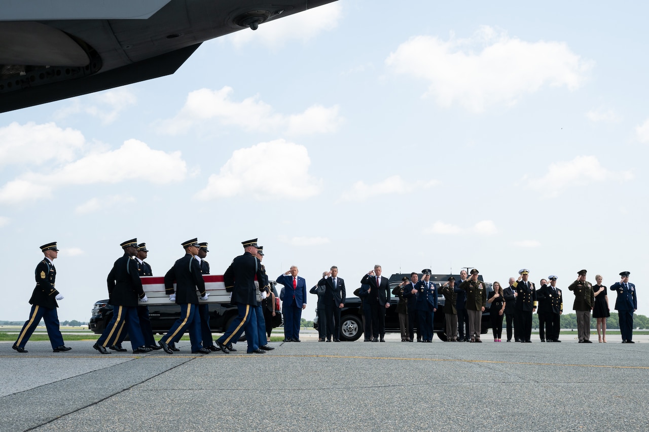 Six men in formal military uniforms carry a flag-draped case as people in similar attire salute in the background.