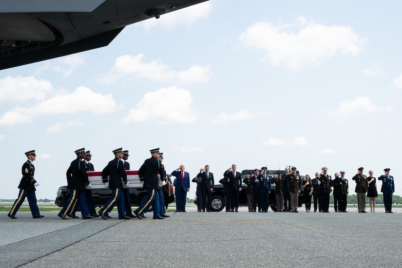 Six men in formal military uniforms carry a flag-draped case as people in similar attire salute in the background.