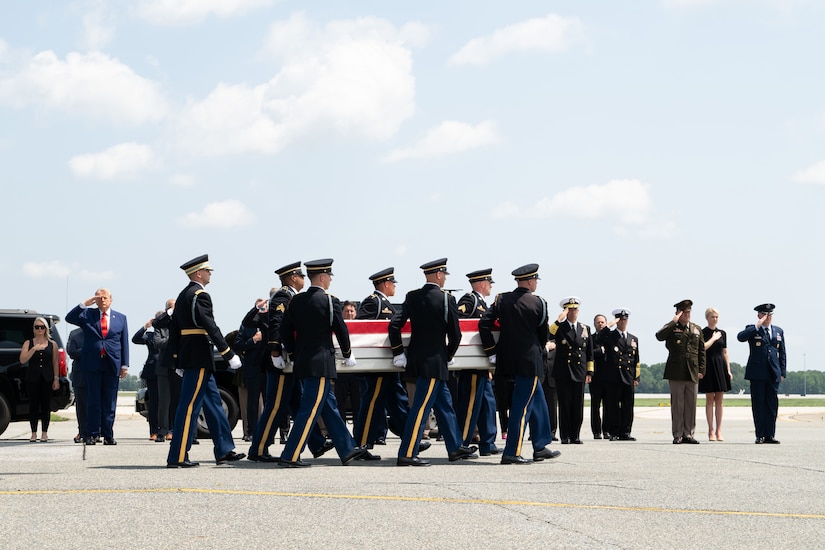 Six men in formal military uniforms carry a flag-draped case as people in similar attire salute in the background.