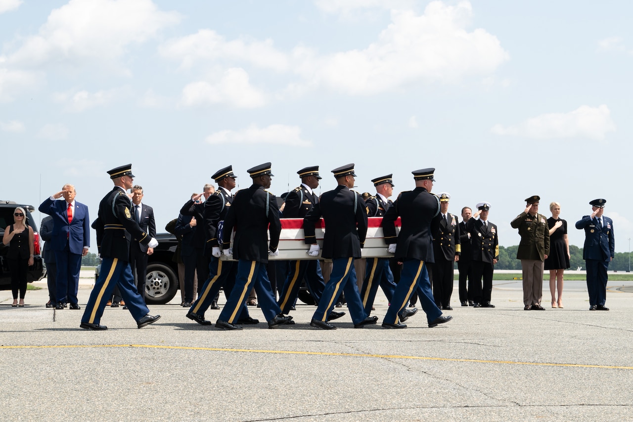 Six men in formal military uniforms carry a flag-draped case as people in similar attire salute in the background.