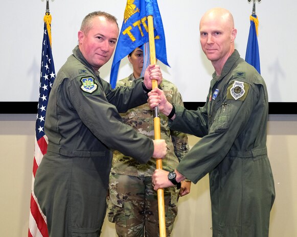 photo of three US Air Force Airman standing on a stage, two Airman are holding a unit flag.