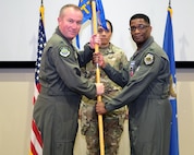 photo of three US Air Force Airman standing on a stage, two Airman are holding a unit flag.