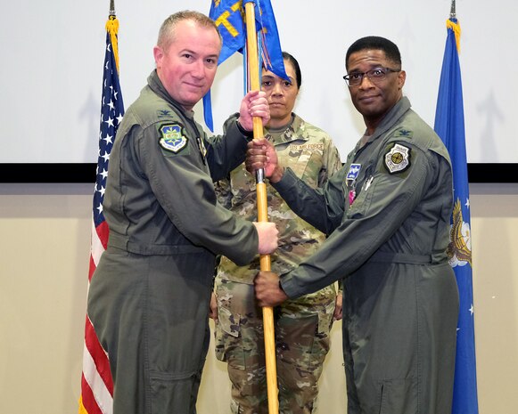 photo of three US Air Force Airman standing on a stage, two Airman are holding a unit flag.