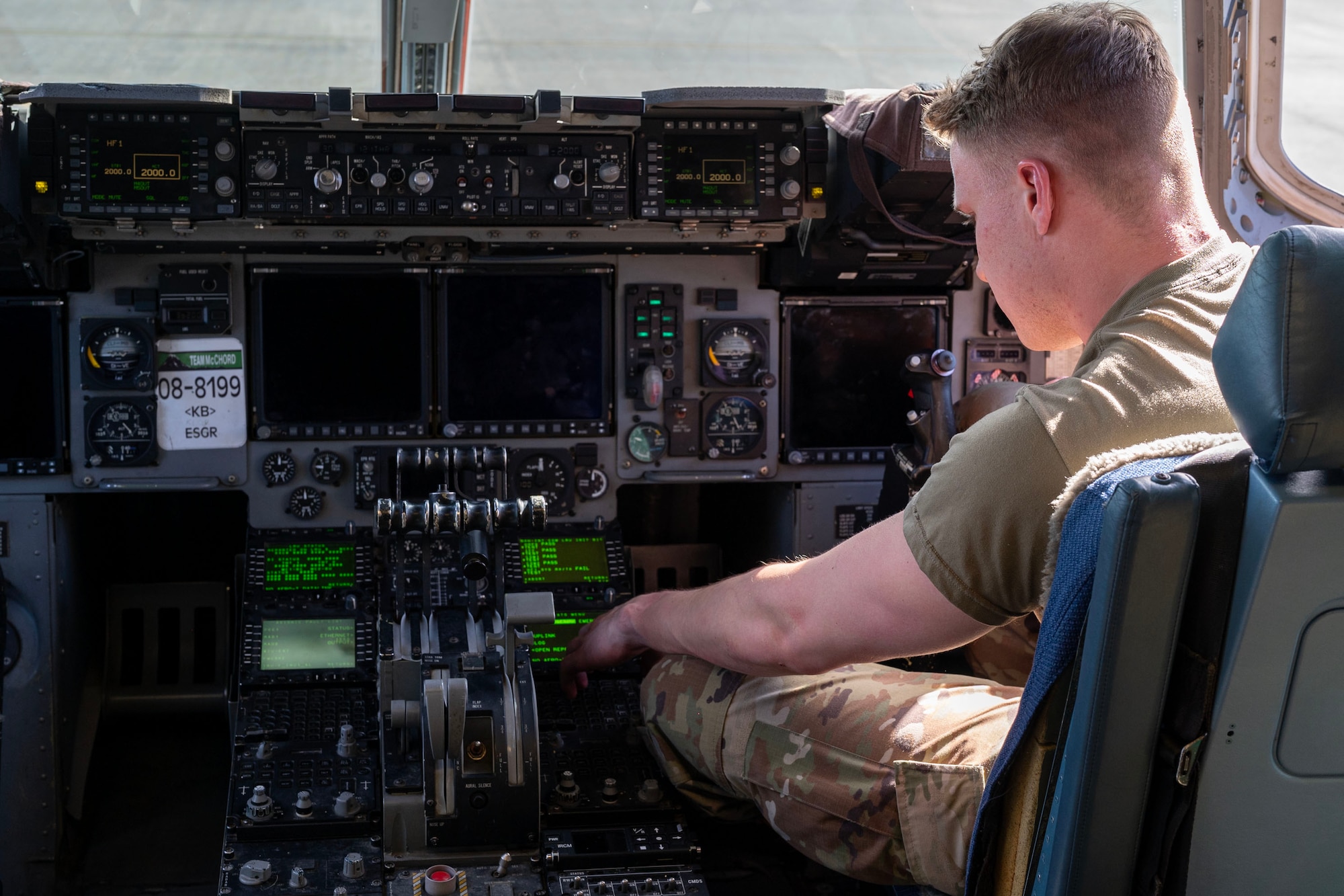 an airman sitting in a chair within an aircraft cockpit, the right right side of the frame shows the airmen, and the left side of the frame shows the panel of switches within the cockpit