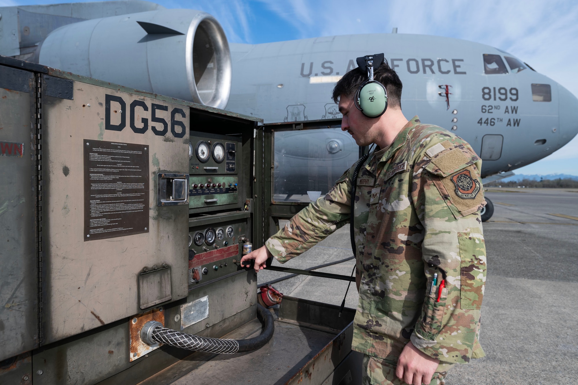 an airman in the right side of the frame, waist up, flips a switch on a generatorr on the left side of the frame. There's a military plane in the background from the side
