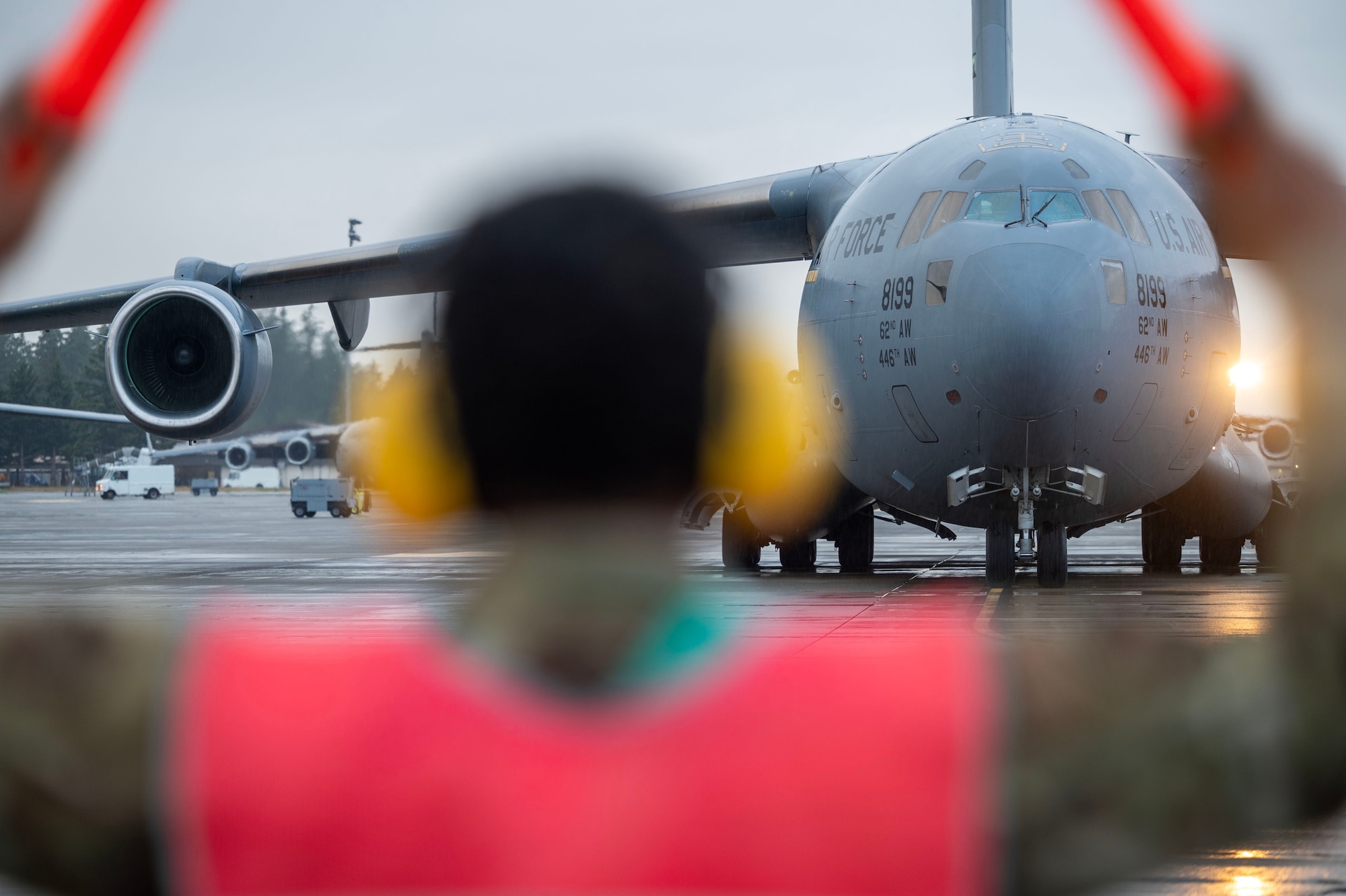 an official in a military uniform and safety vest raises his arms in the foreground of the image. He is blurry. In the background of the image is a military plane